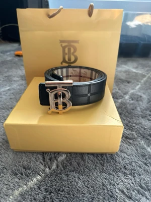 B bälte - Snyggt bälte med klassiskt rutigt mönster och stor metallspänne med B-logga. Är både svart och beige med detaljer i guld. Perfekt accessoar för att lyfta din outfit.