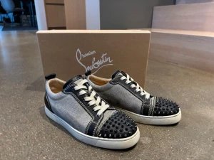 Christian Louboutin sneakers med nitar - Snygga sneakers från Christian Louboutin i grått tyg med svarta läderdetaljer och vita sömmar. Tåpartiet är dekorerat med svarta nitar för en edgy look. Skorna har vita snören och en vit sula. Perfekta för dig som vill sticka ut med din stil.