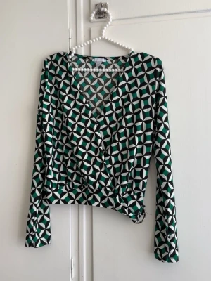 Mönstrad omlottopp från Gina Tricot - Snygg omlottopp från Gina Tricot med grafiskt mönster i grönt, svart och vitt. Toppen har lång ärm, v-ringning och knytdetalj i midjan. Perfekt för dig som gillar färg och mönster!
