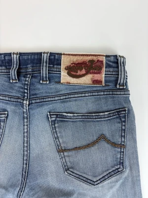 Ljusblå jeans från Jacob Cohen - Snygga ljusblå jeans från Jacob Cohen med klassisk femficksdesign och diskret slitning. Jeansen har raka ben, bruna kontrastsömmar och ett unikt patch med logga bak i midjan. Perfekta för en avslappnad och trendig look.