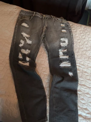 Grå slitna jeans med hål - Säljer ett par grå jeans med kraftiga slitningar och hål på båda benen. Jeansen har klassisk femficksmodell, dragkedjegylf och raka ben. w36 L34