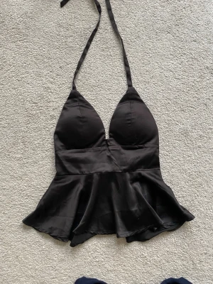 Svart halterneck topp i satin - Snygg svart halterneck topp i glansigt satinmaterial med volang nertill. Toppen har djup v-ringning och knytband i nacken för en justerbar passform. Perfekt för att skapa en trendig och chic look.