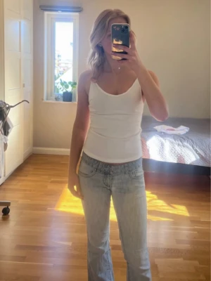 Vit enkel topp med smala band - Säljer en stilren vit topp med smala axelband och rak passform. Den är v ringad framtill och har en lite djupare rygg, men räcker ändå bh bandet. Toppen är ärmlös och har en mjuk, stretchig känsla som gör den perfekt till sommaren. Passar till jeans eller kjol för en clean look.