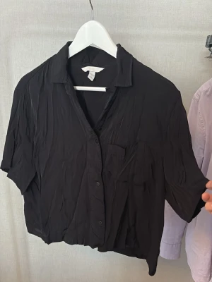 Svart kortärmad skjorta - Svart skjorta med korta ärmar och avslappnad passform. Skjortan har klassisk krage, knäppning framtill och en bröstficka. Perfekt för en chill och stilren look.