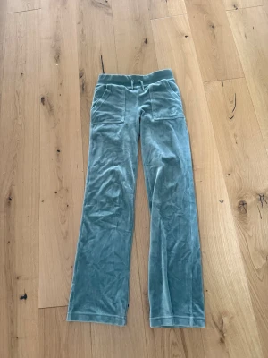 Gröna velourmjukisbyxor Juicy Couture - Mysiga gröna mjukisbyxor i från Juicy Couture med raka ben, bred resår i midjan. Byxorna har två stora fickor framtill och en broderad JC-logga på ena fickan. Perfekta för en chill och trendig look.