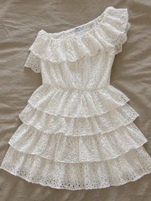 Vit volangklänning från Loavies - Superfin vit klänning från Loavies med volanger i flera lager och ett snyggt broderat spetsmönster. Klänningen har en offshoulder-design och kort ärm på ena sidan, vilket ger en unik och trendig look🩷🩷