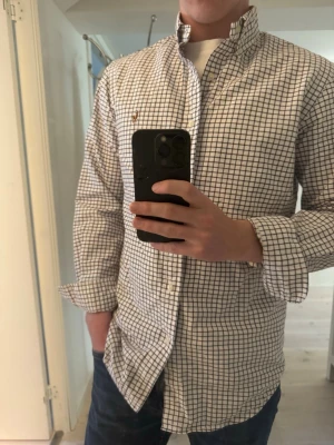 Rutig skjorta från Ralph Lauren - Klassisk vit skjorta med mörkblå och bruna rutor från Ralph Lauren. Skjortan har knappar framtill, button-down krage och lång ärm. Tillverkad i bomull för en skön och avslappnad känsla. Perfekt till jeans eller chinos.