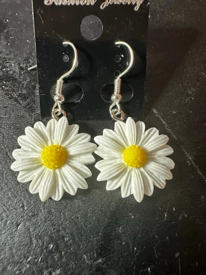 Örhängen med prästkrageblommor - Superfina örhängen i form av prästkragar med vita kronblad och gul mitt. Hängande modell med silvriga krokar. Perfekta för dig som vill ha en söt och lekfull accessoar med blommig känsla. Materialet är troligtvis plast och metall.