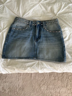 Blå jeanskjol med nitar - Snygg blå jeanskjol med silvriga nitar längs midjan och fickorna. Klassisk femficksmodell med dragkedja och knapp framtill. Kjolen har en kort passform och är perfekt för dig som gillar en edgy stil. Perfekt för sommar