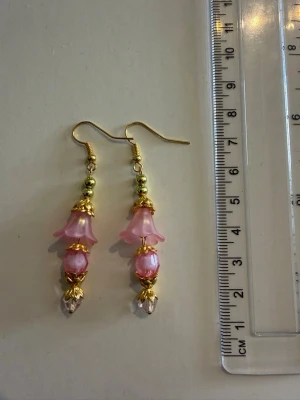 Rosa blommiga örhängen med pärlor - Säljer ett par handgjorda örhängen med rosa blommor i plast, guldfärgade detaljer och pärlliknande dekorationer. Örhängena har en hängande design och är ca 6 cm långa. Perfekta för dig som gillar unika accessoarer med romantisk känsla. Nickelfria 