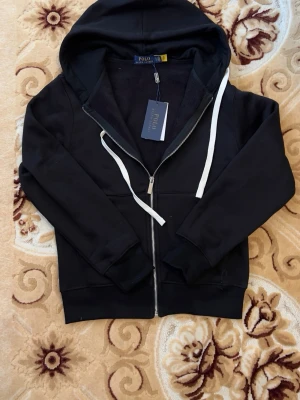 Svart hoodie från Polo Ralph Lauren - Svart hoodie från Polo Ralph Lauren med dragkedja och vita snören i huvan. Tillverkad i mjukt material med fleecefoder på insidan. Klassisk design med ribbade muddar och fickor framtill. Perfekt för en chill och stilren look.