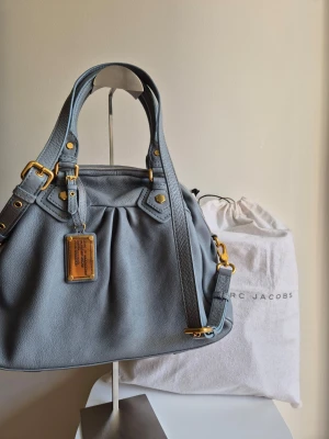 Ljusblå handväska från Marc Jacobs Classic Q  - Snygg ljusblå handväska från Marc Jacobs i mjukt läder med guldfärgade metalldetaljer. Väskan har dubbla handtag, avtagbar axelrem och dekorativ metallbricka framtill. Perfekt för dig som vill ha en stilren och trendig accessoar. Det kan hämtas i Solna/t-centralen eller frakt 