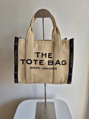 Beige Tote bag från Marc Jacobs - Trendig beige shoppingväska från Marc Jacobs i canvas med svarta detaljer och tydlig logga framtill. Väskan har dubbla handtag och svarta sidor med vita tryck. Perfekt för dig som vill ha en snygg och rymlig tote bag med streetkänsla. Endast en detalj det saknas en lång axelrem. 