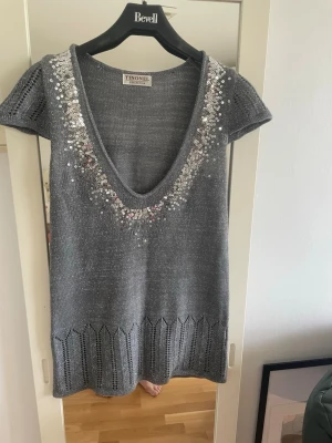 Grå stickad topp med paljetter Tinonel - Grå stickad topp från Tinonel Collection med djup v-ringning och korta ärmar. Utsmyckad med silvriga och vita paljetter längs ringningen. Tunt, glansigt material och dekorativt mönster nertill. Perfekt för dig som gillar att sticka ut.