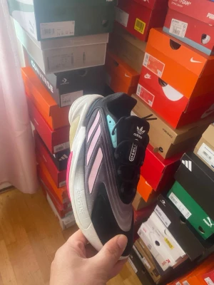 Adidas Ozelia svart/lila sneakers - Snygga Adidas Ozelia sneakers med svart ovandel, lila och rosa detaljer samt vit sula. Skon har klassiska tre ränder på sidan och en modern, chunky siluett. Materialet är en mix av mocka och syntet. Perfekt för dig som vill sticka ut med en unik färgkombo.