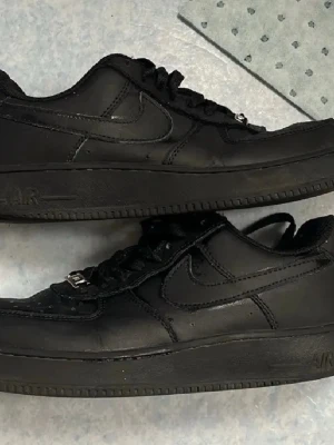 Nike Air Force 1 Low helsvart läder - Klassiska Nike Air Force 1 Low i helsvart läder med svart sula och svarta skosnören. Swoosh-logga på sidorna och AIR-detalj på sulan. Perfekta sneakers för en clean och stilren look.