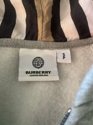 Grå hoodie från Burberry med dragkedja - Snygg grå hoodie från Burberry med dragkedja och klassiskt mönstrat foder i huvan. Hoodien har fickor framtill och justerbar dragsko vid huvan. Perfekt för en stilren och avslappnad look.