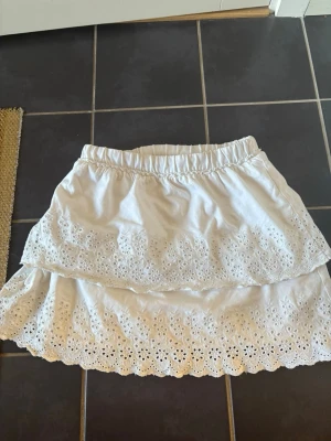 Vit volangkjol med broderi från Zara - Superfin vit kjol från Zara med volanger och broderade spetsdetaljer. Kjolen har resår i midjan och är perfekt för en somrig look. Tillverkad i bomull för en skön och luftig känsla.