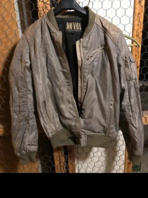 Grå/grön bomberjacka från Jofama - Snygg grå/grön bomberjacka från Jofama med klassisk passform, ribbade muddar och krage i olivgrönt. Jackan har flera fickor med dragkedja, inklusive en ärmficka. Perfekt för dig som gillar streetwear och vill ha en cool look.