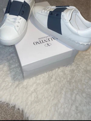 Valentino Garavani vita sneakers - Snygga vita sneakers från Valentino Garavani med bred mörkblå detalj över snörningen och klassisk logga på tungan. Skorna har en låg profil och är tillverkade i läder med gummisula och nitar på hälen. Perfekt för dig som vill ha en exklusiv look.