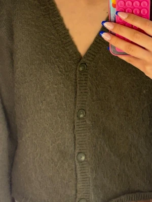 Mörkgrön stickad kofta med knappar - !!DEN SKA VARA NOPPRIG!! Mysig mörkgrön stickad kofta med v-ringning och stora knappar framtill. Koftan har en fluffig textur och ribbade kanter vid ärmslut och längs knappraden. Perfekt att slänga på sig när det är kyligt ute. Säljer för att jag inte använder den , använd 1 gång