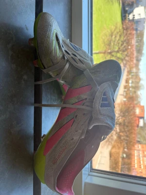 Adidas Predator fotbollsskor grå/rosa - Säljer ett par Adidas Predator fotbollsskor i grått med rosa och limegröna detaljer. Skorna har klassisk snörning, täckt plös och mönstrad ovandel för bättre bollkontroll. Snygg design med Adidas-logga på plösen och sidorna. Perfekta för dig som vill sticka ut på planen!