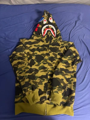 BAPE camo hoodie med shark-motiv - Säljer en grön camo hoodie från BAPE med ikoniskt shark-motiv på huvan och broderade detaljer. Hoodien har dragkedja, ribbade muddar och coola färgglada patchar på huvan. Perfekt för dig som vill sticka ut och gillar streetwear.