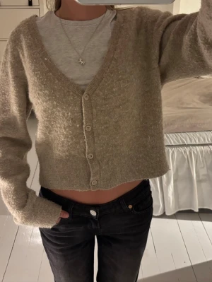 Beige cropped kofta med v-ringning - Mysig beige kofta med v-ringning och fyra knappar framtill. Modellen är cropped och har långa ärmar. Perfekt att slänga över en t-shirt för en avslappnad look. Koftan är stickad i ett mjukt material och har en enkel, stilren design.