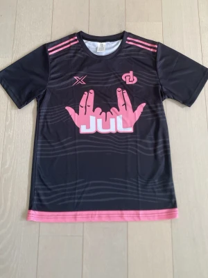 Jul tröja - Snygg svart sporttröja med rosa tryck och detaljer. Framsidan har ett stort rosa handmotiv och JUL-logga, samt två mindre loggor på bröstet. Tröjan har rund hals, korta ärmar och rosa ränder på axlarna. Tillverkad i ett lätt och snabbtorkande material.