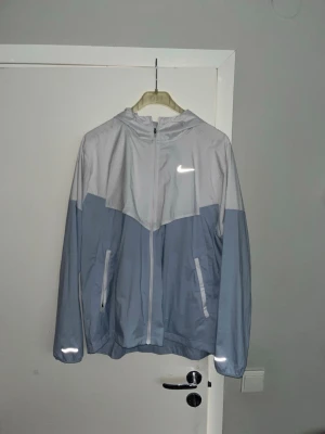Nike vindjacka i ljusblå och vit - Nike vindjacka med huva i snygg tvåfärgad design, ljusblå nedtill och vit upptill. Jackan har dragkedja framtill, två fickor med dragkedja och reflekterande detaljer. Perfekt för blåsiga dagar och sportiga outfits.