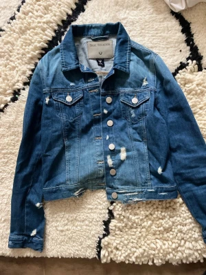  True Religion - Säljer en blå jeansjacka från True Religion med slitna detaljer och silverfärgade knappar. Klassisk modell med två bröstfickor och krage. Jackan har en cool, avslappnad vibe och är tillverkad i jeansmaterial.
