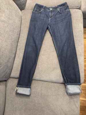 Blå jeans från Burberry brit - Ett par mörkblå skinny jeans från Burberry Brit med låg midja (low rise). Jeansen har en stilren och modern passform som sitter tajt längs benen. De är designade med fem fickor, bälteshällor och stängs med dragkedja och knapp. Synliga sömmar och en läderpatch med Burberry Brit-logotypen baktill ger en exklusiv känsla. Uppvikta benslut avslöjar Burberrys ikoniska rutmönster på insidan, vilket ger en snygg och unik detalj.  Observera att modellen är liten i storleken.