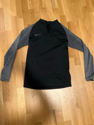 Svart träningsöverdel från Nike - Svart långärmad träningsöverdel från Nike med gråmelerade ärmar och halv dragkedja framtill. Tillverkad i ett stretchigt funktionsmaterial som andas, perfekt för träning eller uppvärmning. Nike-logga på bröstet ger en sportig look.