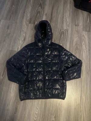Mörkblå pufferjacka från H&M - Snygg mörkblå pufferjacka från H&M med glansig finish och quiltad design. Jackan har huva, dragkedja framtill och är vadderad för extra värme. Perfekt för kalla dagar när du vill hålla stilen och samtidigt vara varm.