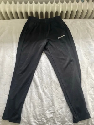 Svarta Nike Dri-FIT mjukisbyxor - Säljer ett par svarta mjukisbyxor från Nike med Dri-FIT-material som andas och håller dig torr. Byxorna har en smal passform, resår i midjan och en vit Nike-logga på låret. Perfekta för träning eller chill dagar.