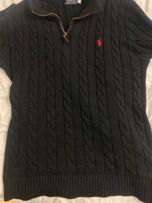 Mörkblå kabelstickad tröja Polo Ralph Lauren - Kabelstickad mörkblå tröja från Polo Ralph Lauren med halv dragkedja och röd broderad logga på bröstet. Tröjan har ribbade muddar och krage, samt ett klassiskt och tidlöst utseende. Perfekt för lager på lager och snygg till jeans.