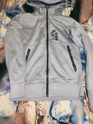 Nike Air Max ljusblå hoodie - Nike Air Max hoodie i ljusblått med dragkedja framtill, två snedställda fickor med svarta dragkedjor och tryckt logga på bröstet. Hoodien har huva och breda muddar vid ärmslut och nederkant. Perfekt för en sportig och avslappnad stil.