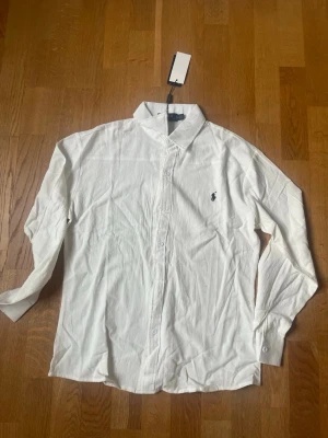 Vit skjorta från Polo Ralph Lauren - Säljer en klassisk vit skjorta från Polo Ralph Lauren med broderad logga på bröstet. Skjortan har långärmad design, knappar framtill och är tillverkad i bomull. Perfekt för dig som gillar stilrena plagg med ikonisk branding.