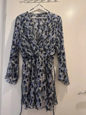Blommig omlottklänning från Zara - Snygg kort omlottklänning från Zara med blått och svart blommönster på vit botten. Klänningen har långa ärmar, knyt i midjan och lätt volangkant nertill. Perfekt för dig som gillar en feminin och trendig stil.