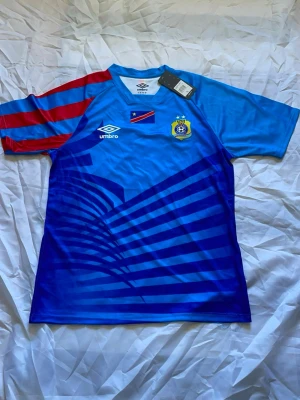 Blå Umbro fotbollströja DR Kongo - Snygg blå fotbollströja från Umbro med DR Kongos landslagsemblem och flagga på bröstet. Ena ärmen har röda och blå ränder, medan resten av tröjan har grafiskt mönster i olika blå nyanser. Tillverkad i lätt och ventilerande material, perfekt för träning eller match.