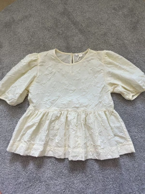 Vit blus med puffärm från Gina Tricot - Säljer en vit blus från Gina Tricot med puffiga korta ärmar och strukturerat tyg. Blusen har rund halsringning och en volang nedtill som ger en söt och trendig look. Perfekt för dig som gillar stilrena och feminina plagg.