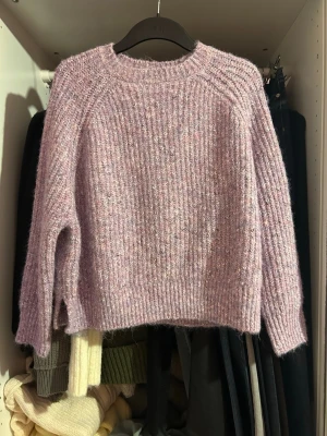 Ljuslila stickad tröja Urban Outfitters - Mysig ljuslila stickad tröja från Urban Outfitters. Tröjan har en lös passform, rund halsringning och ribbstickade muddar. Garnet är fluffigt och har inslag av vitt och rosa, vilket ger en mjuk och trendig look.