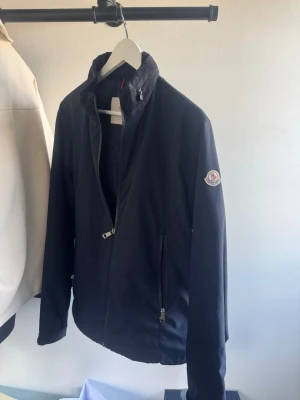 Mörkblå vindjacka från Moncler - Snygg mörkblå vindjacka från Moncler med hög krage och dragkedja framtill. Jackan har två fickor med dragkedja och Moncler-logga på ärmen. Tillverkad i ett lätt och vindtätt material, perfekt för blåsiga dagar.