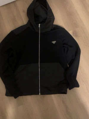 Prada jacka - Snygg svart hoodie från Prada med dragkedja framtill och klassisk huva. Hoodien har ett diskret Prada-logo på bröstet och är tillverkad i ett mjukt material som känns bekvämt. Perfekt för en stilren och avslappnad look.