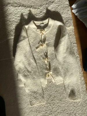 Krämvit kofta med rosettdetaljer - Superfin krämvit kofta med fyra söta rosetter framtill istället för knappar. Koftan är stickad i ett mjukt, fluffigt material och har långa ärmar samt rund halsringning. Perfekt för lager-på-lager och ger en romantisk touch till din outfit.