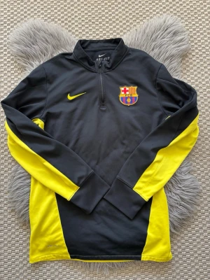 Nike Barcelona Dri-Fit tröja svart/gul - Snygg långärmad träningströja från Nike med FC Barcelona-logga på bröstet. Tröjan är svart med gula detaljer och har halv dragkedja framtill. Tillverkad i Dri-Fit-material som andas och passar perfekt för sportiga dagar.