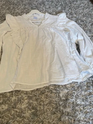 Vit blus med volangdetaljer - Söt vit blus med volangdetaljer längs axlarna och bröstet. Blusen har en lös passform, V-ringning och långa ärmar. Materialet ser ut att vara lätt och luftigt, perfekt för en trendig och avslappnad look.