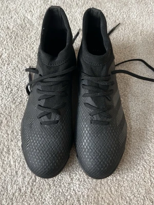 Svarta Adidas fotbollsskor med sockliknande skaft - Säljer ett par helsvarta fotbollsskor från Adidas med sockliknande skaft och snörning. Skorna har en texturerad ovandel för bättre bollkontroll och mönstrad sula som ger bra grepp på konstgräs. Perfekta för dig som vill ha stilrena och funktionella skor på planen.