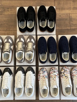 Maison Margiela sneakers i olika färger - Flera par Maison Margiela sneakers i olika färger och designer. Finns i svart, grå/vit, marinblå samt vita modeller med färgstänk. Klassisk låg siluett med snörning och ovandel i mocka och läder. Perfekta för dig som gillar unik och trendig stil.