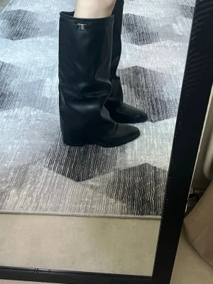 Svarta höga läderstövlar med kilklack - Snygga svarta stövlar i läder med kilklack och högt skaft. Stövlarna har en stilren design med ett diskret spänne upptill. Perfekta för att lyfta din outfit och ge en edgy vibe.